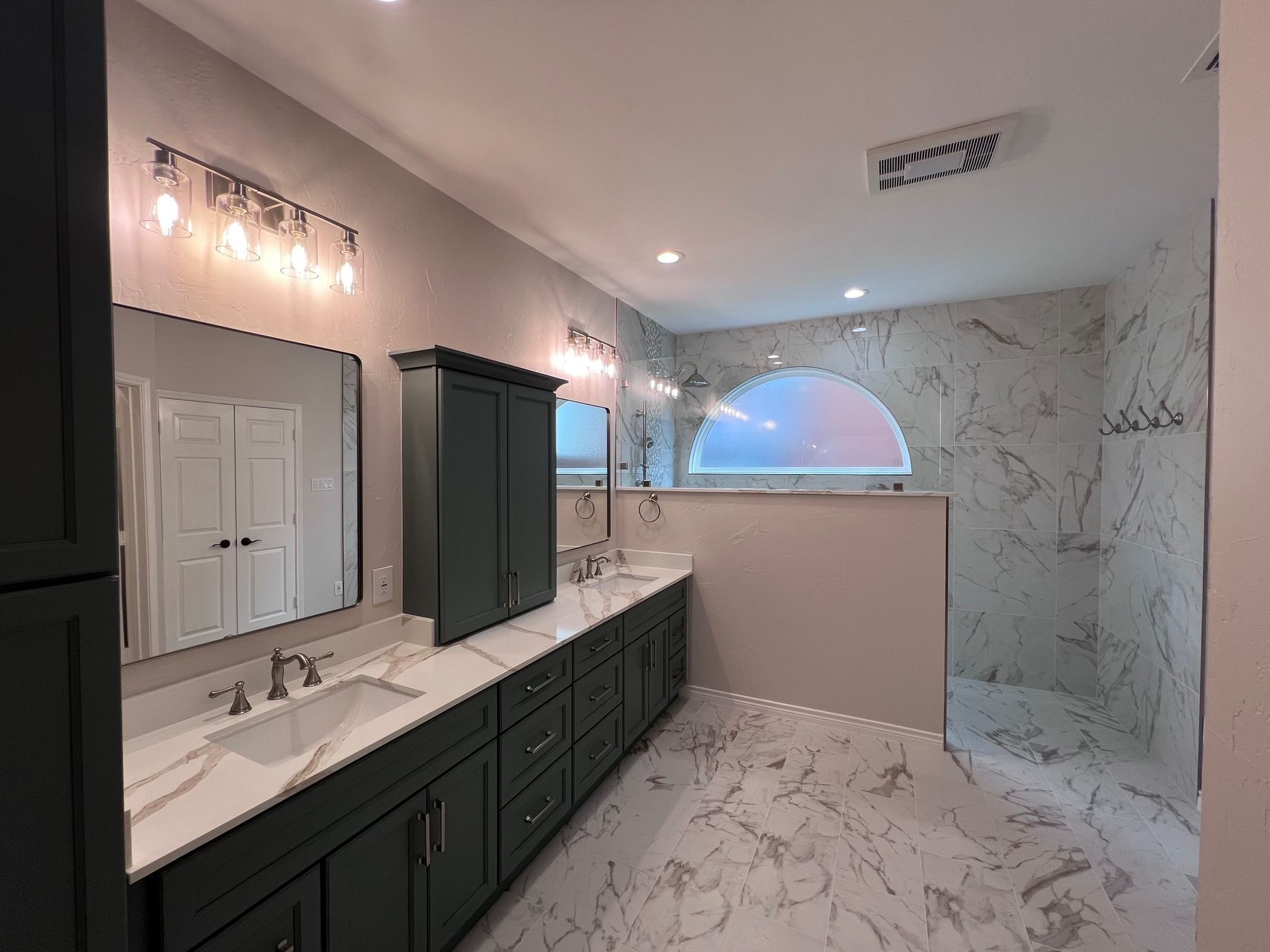 Modern bathroom with dark teal cabinets, marble-look tile, and a large arched window above a walk-in shower.