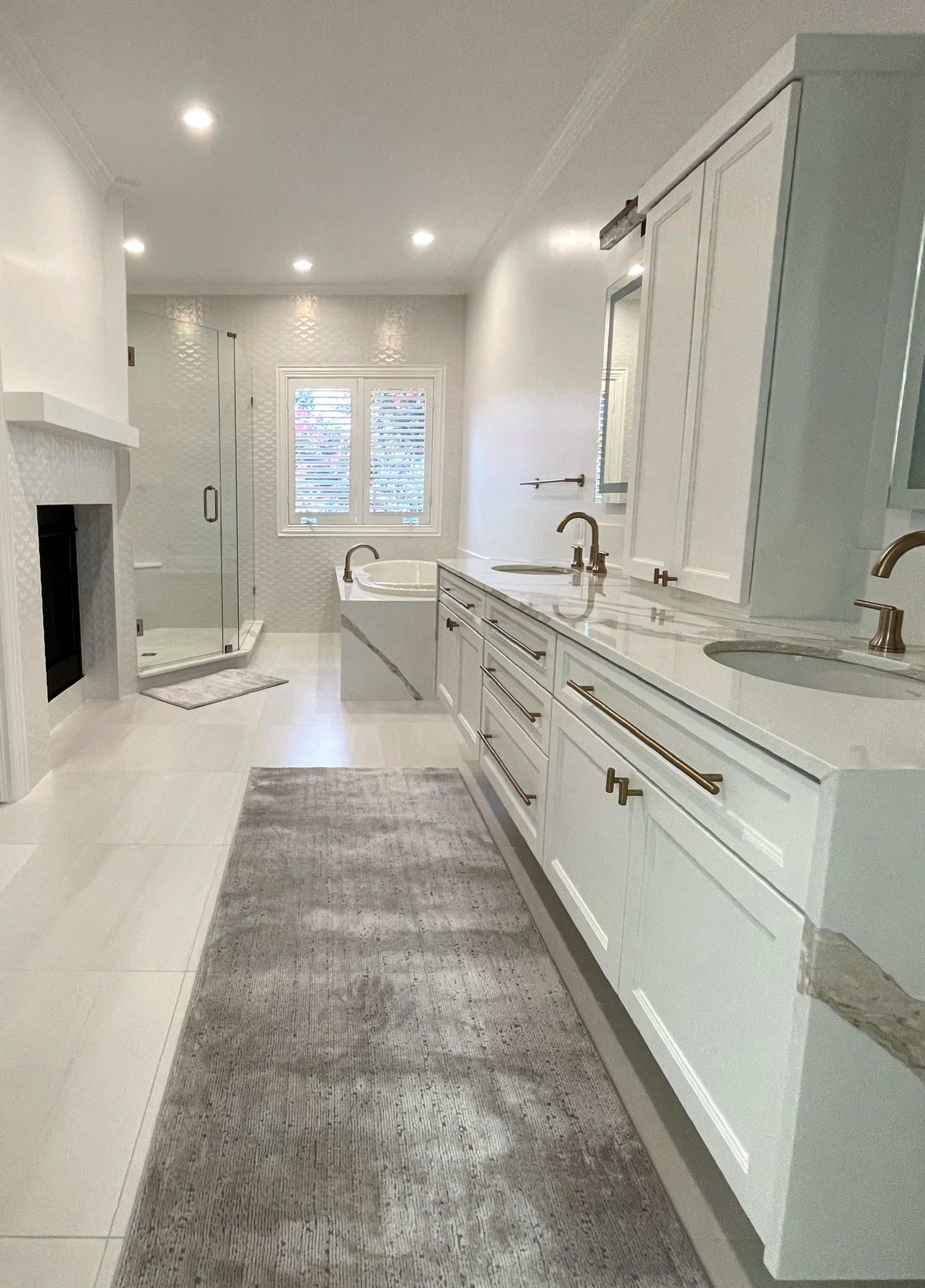 Spacious white bathroom with marble counters, long vanity, large rug, and glass shower.