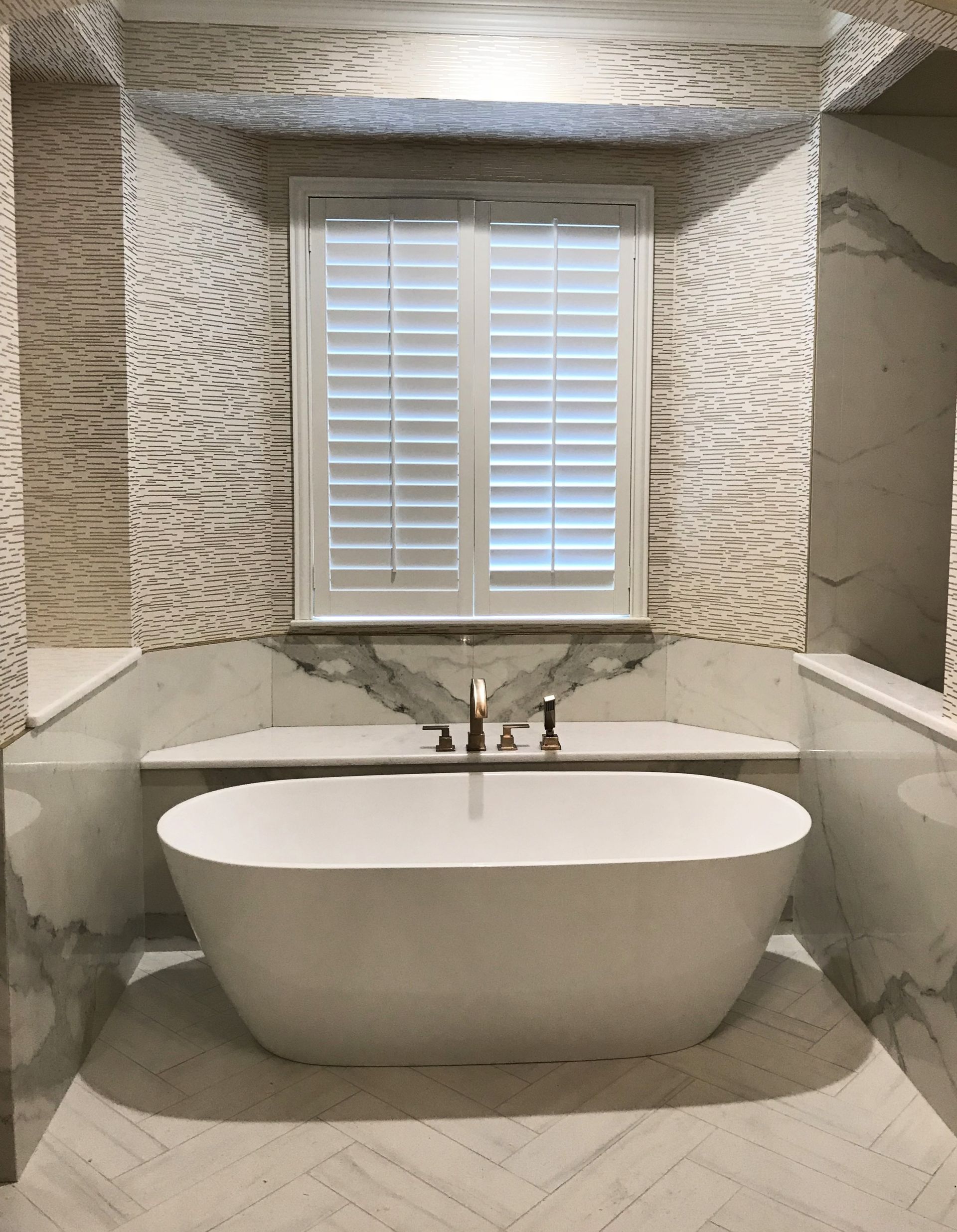 Freestanding white bathtub below a window with closed shutters, set in a marble-tiled bathroom.