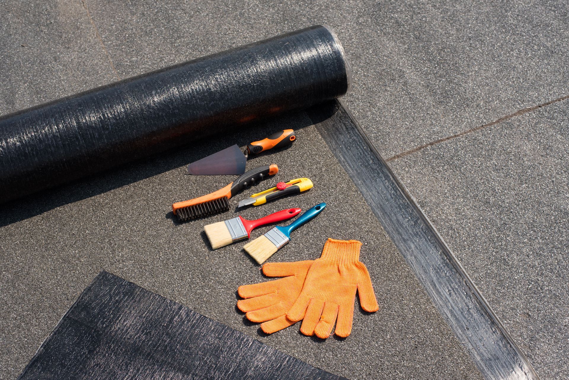 Roll of black roofing material with tools and orange work gloves on a dark roof.