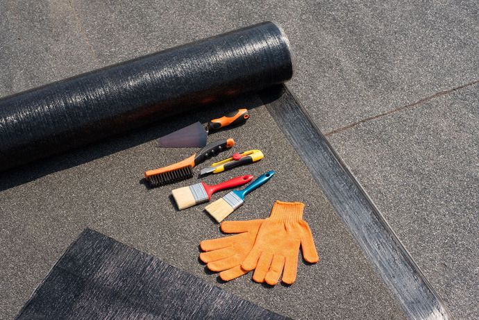Roll of black roofing material with tools and orange work gloves on a dark roof.