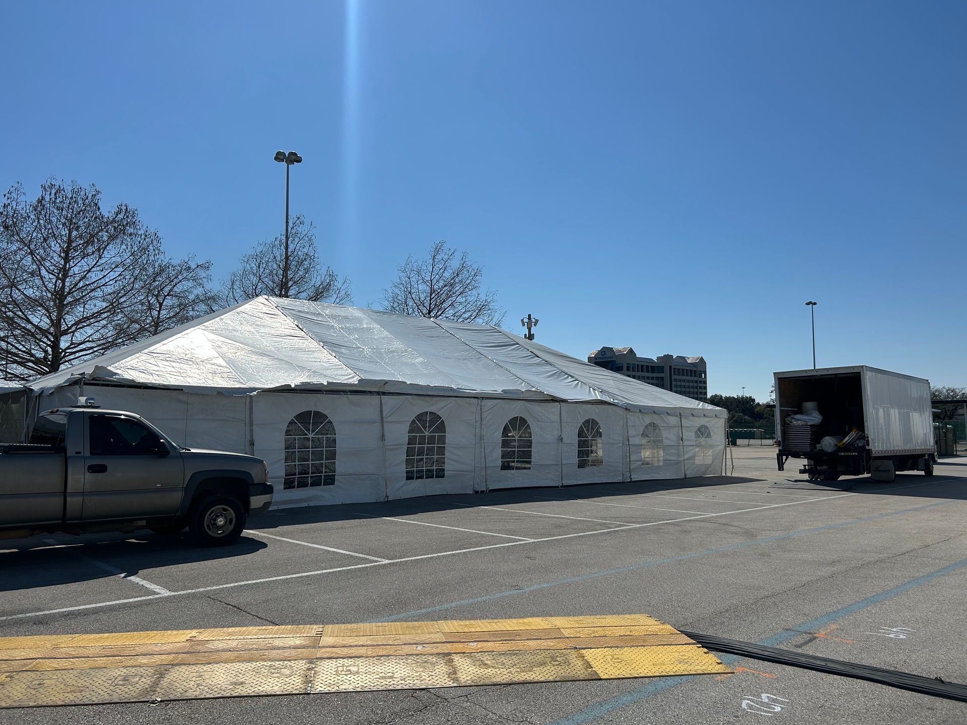 A large white tent set up in a parking lot. A truck and pickup truck are nearby on a sunny day.