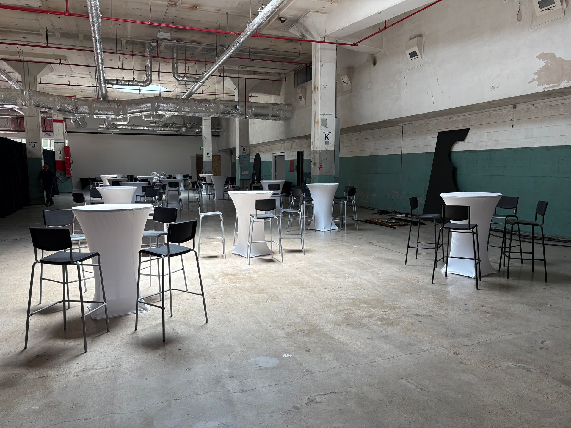 An empty industrial space set up with white high-top tables and black chairs, ready for an event. The walls are white, turquoise, and aged.
