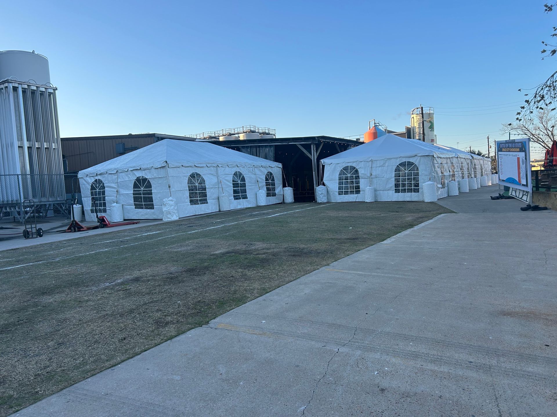 Two white tents with arched windows set up on a grassy area next to a sidewalk. A water tower is visible in the background.