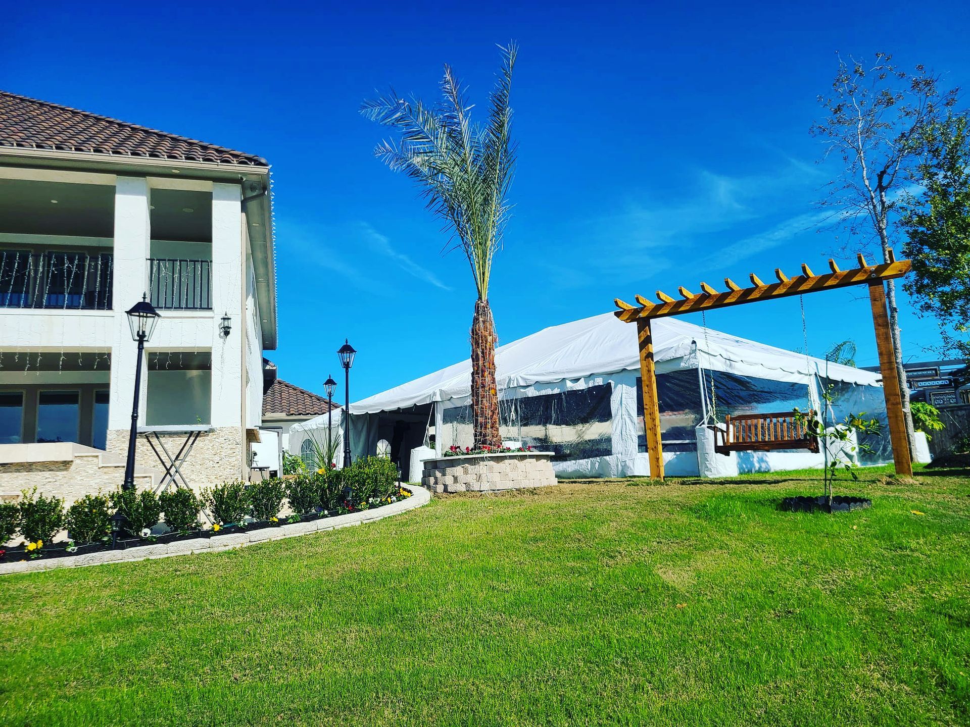 Lawn with white building, clear tent, pergola swing, palm tree, and clear blue sky.