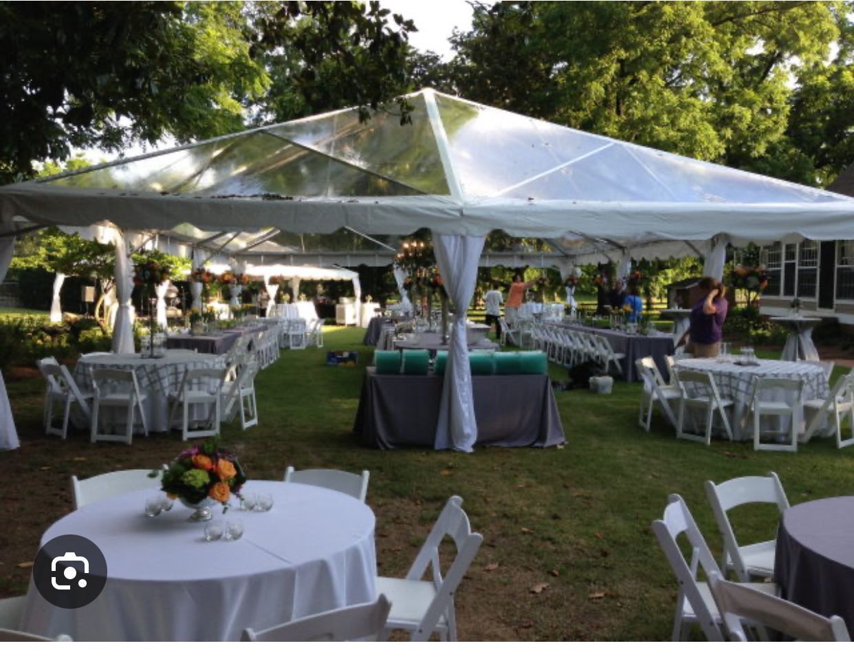 Outdoor event with clear-topped tent, round tables with white linens, and white chairs on a lawn.