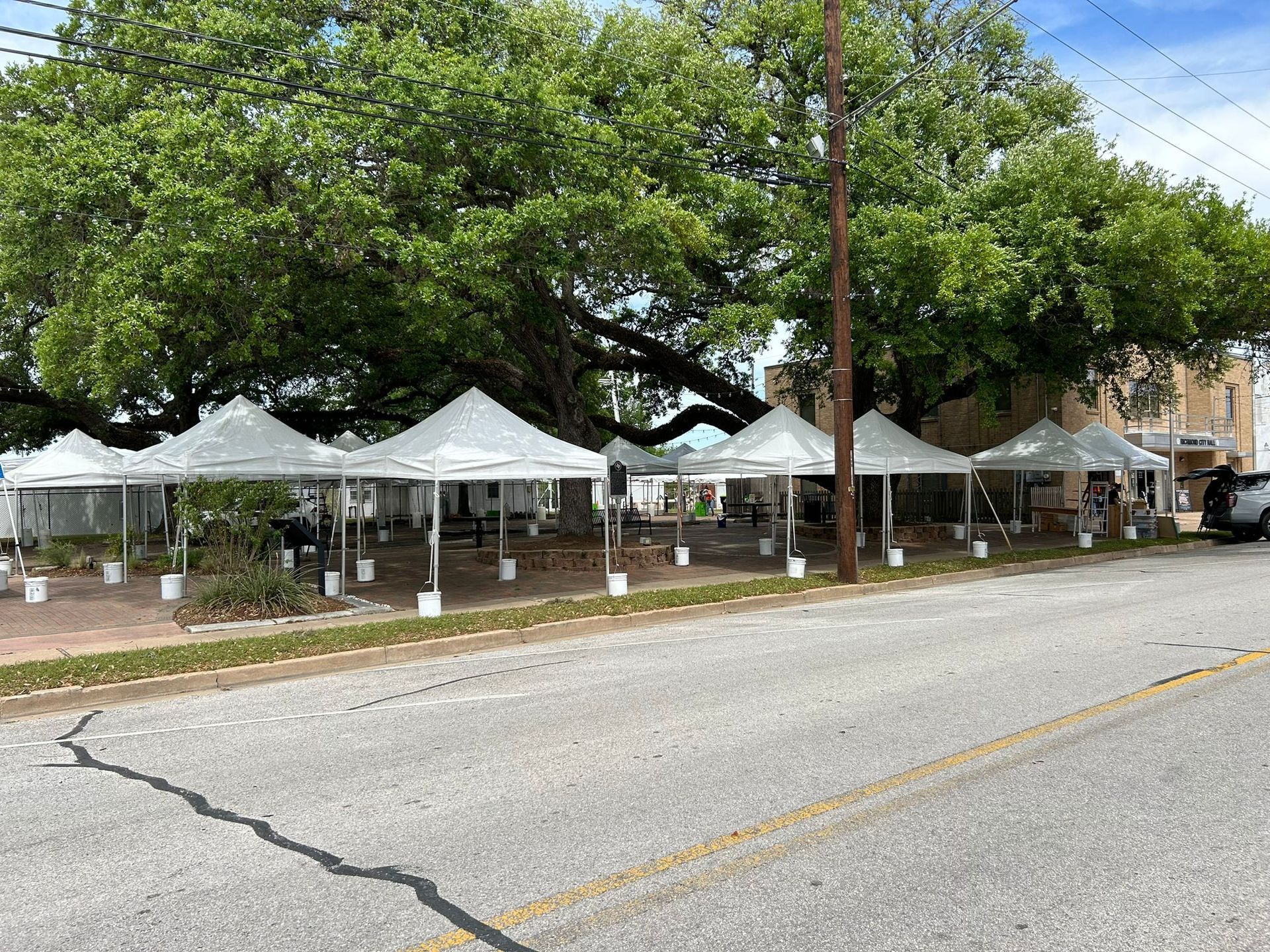 Outdoor dining area with multiple white tents set up on a sidewalk under a large tree.