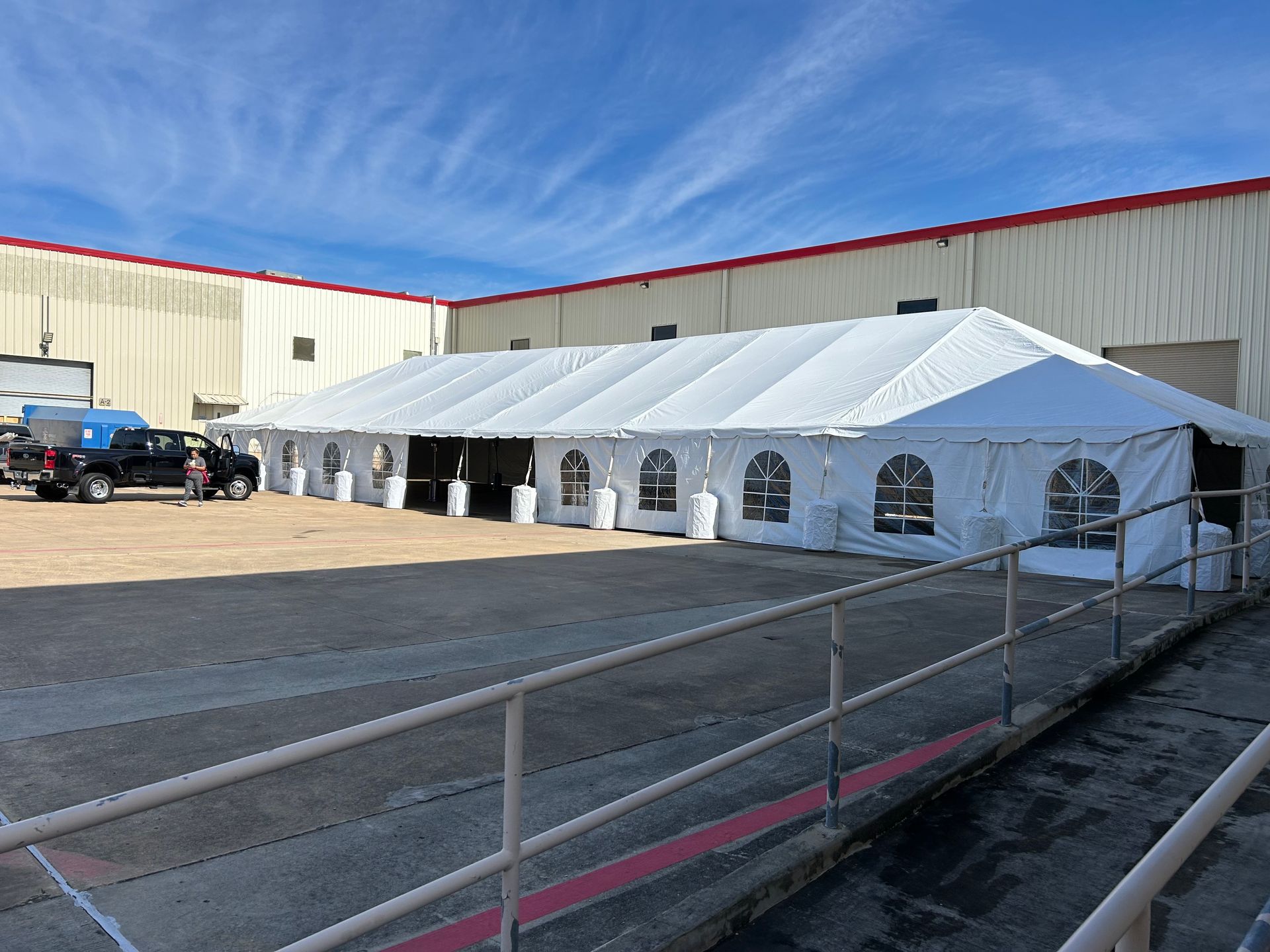 A large white tent set up in a parking area next to a brick building. A black truck is parked nearby.
