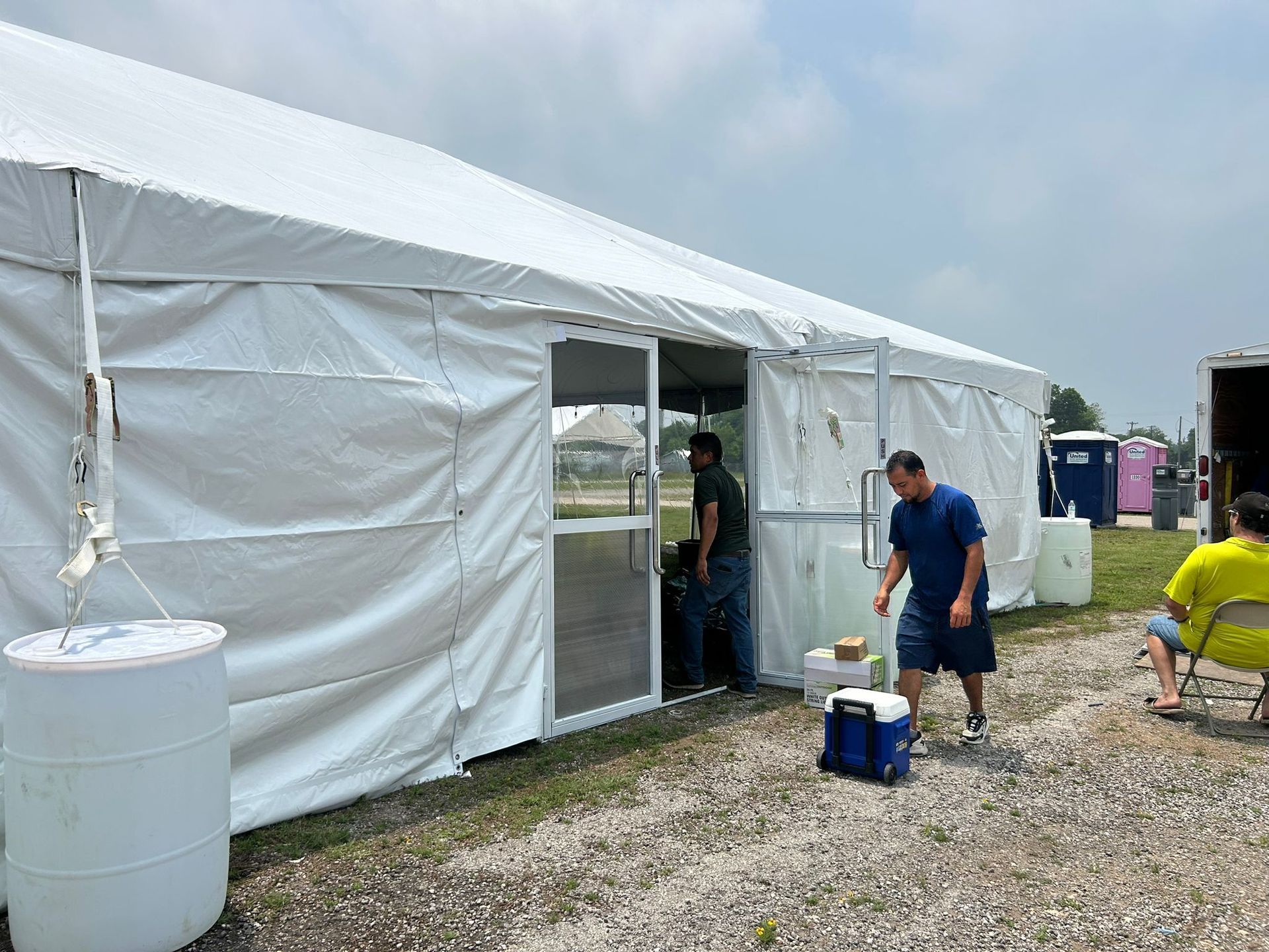 White tent entrance with people entering/exiting. A cooler and water barrels sit outside on gravel.
