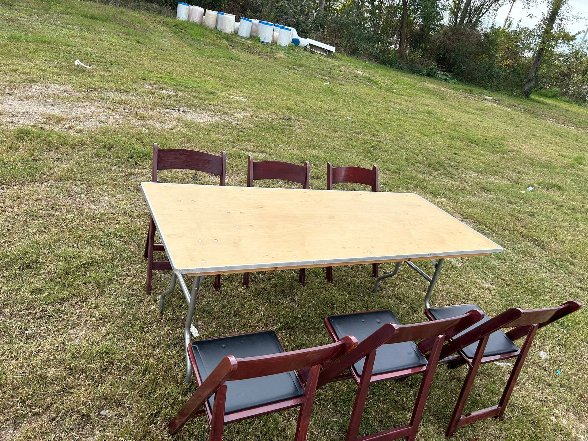 A rectangular table and six folding chairs sit on a grassy hill. White buckets are in the background.