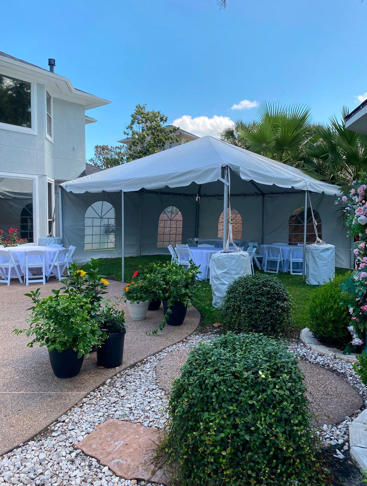 Backyard event with tent, tables, and potted plants. White tent, blue sky, and green lawn.