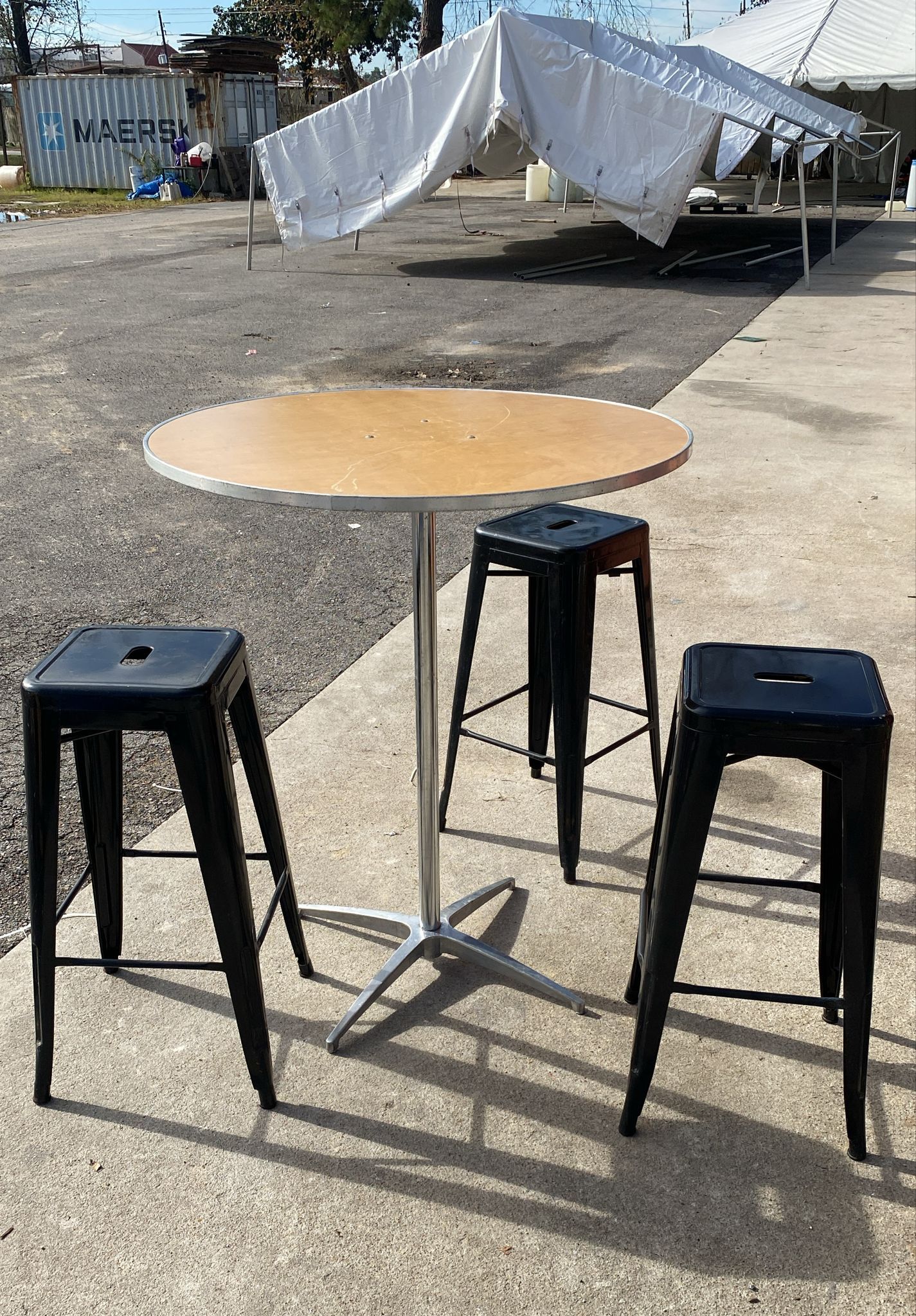 Three black bar stools surround a round wooden table. A white tent is in the background.