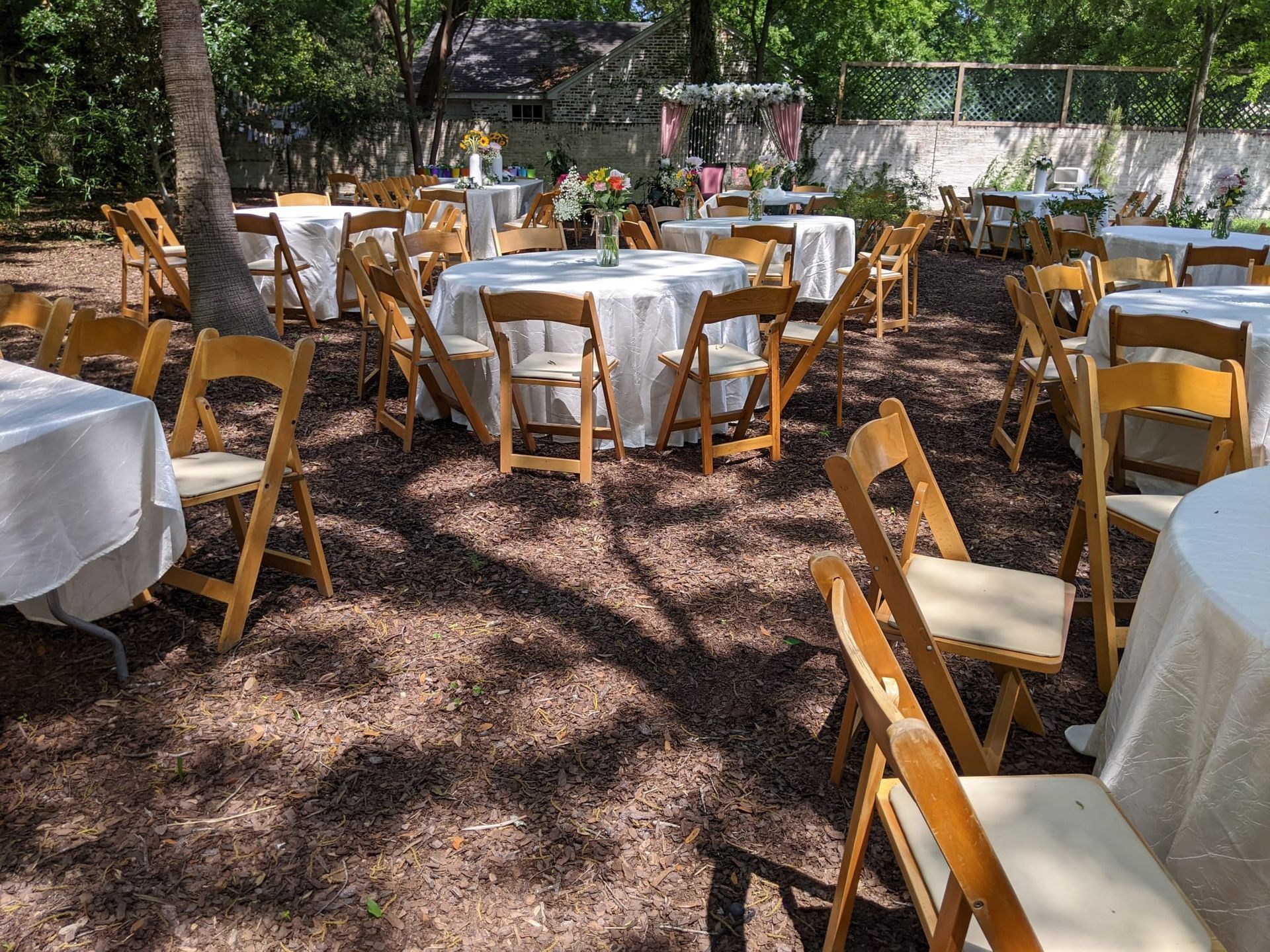 Outdoor event setup with round tables, white tablecloths, wooden chairs, and floral centerpieces.