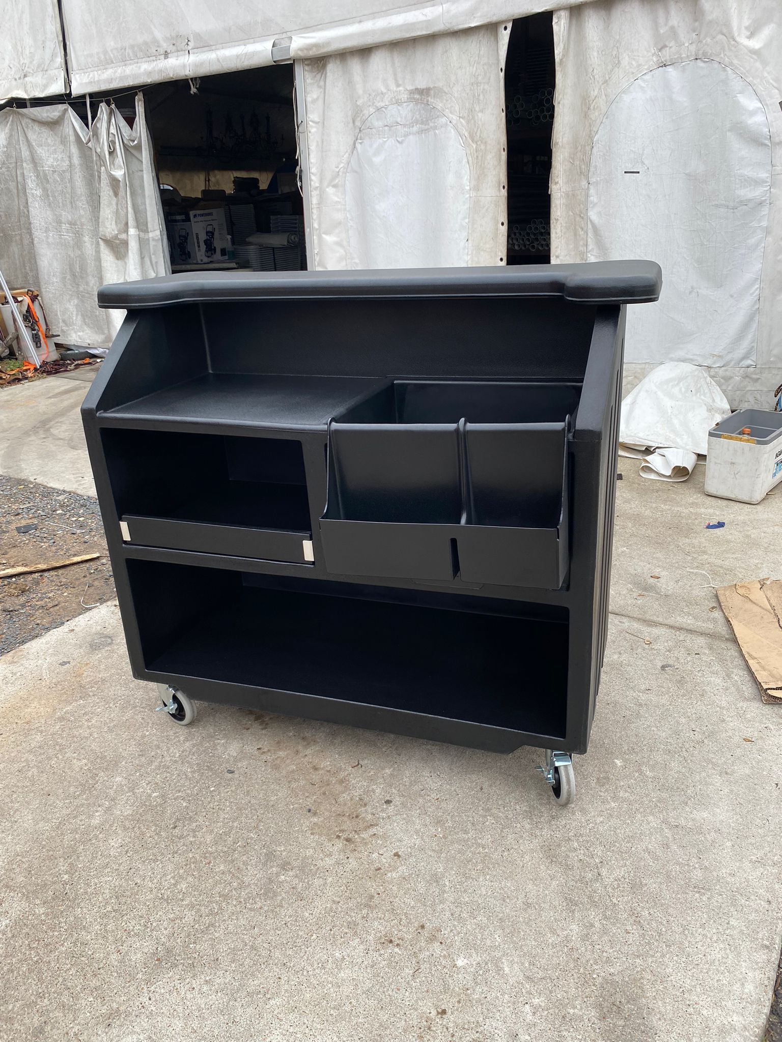 Black portable bar with shelving and compartments on wheels, outdoors.