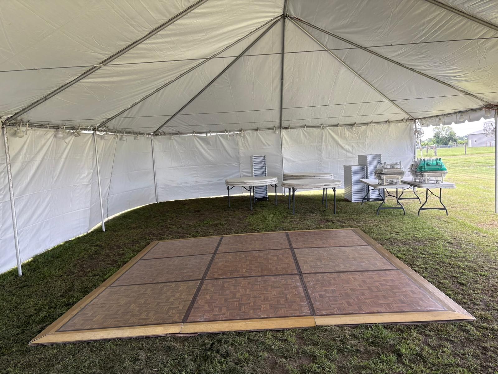 Inside a white tent on grass: a dance floor, tables with stacks of plates, and a food warmer.
