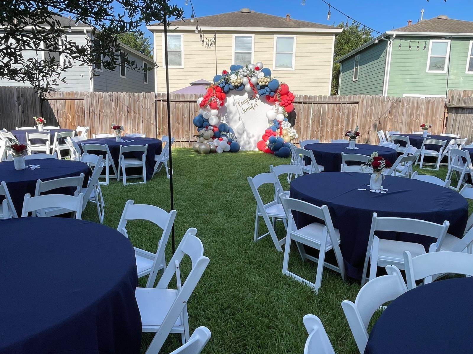 Outdoor party setup: round tables with blue cloths, white chairs, a balloon arch, and string lights.