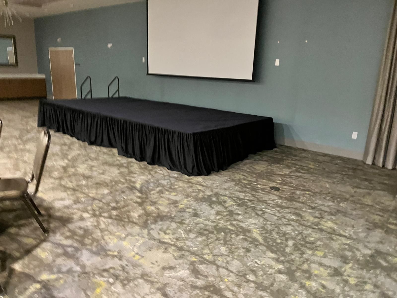A rectangular black stage with black skirting in a room with blue walls, a screen, and patterned carpet.