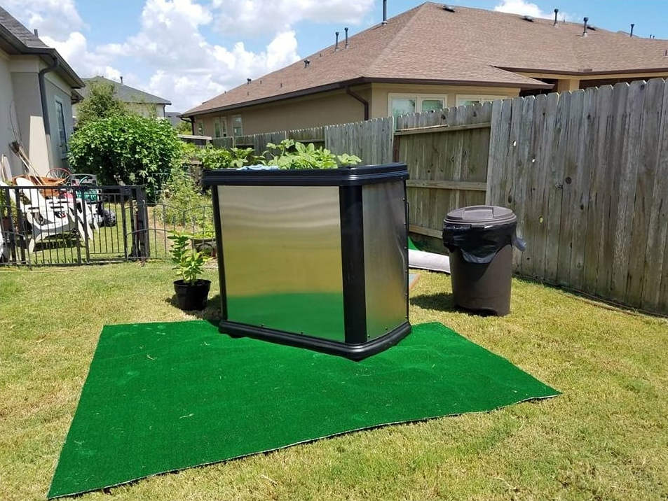 A metal and black container garden on a green mat in a backyard. A trash can sits next to it.