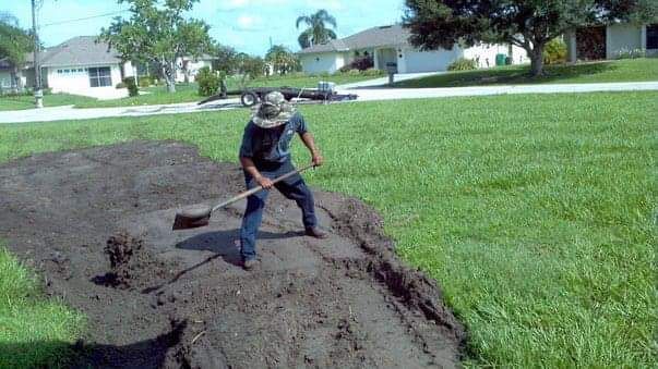 Person shoveling dark material on a grassy lawn; houses in the background.