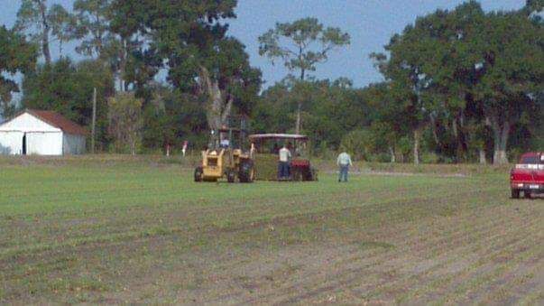 Field with machinery and people, white building and red truck in background.