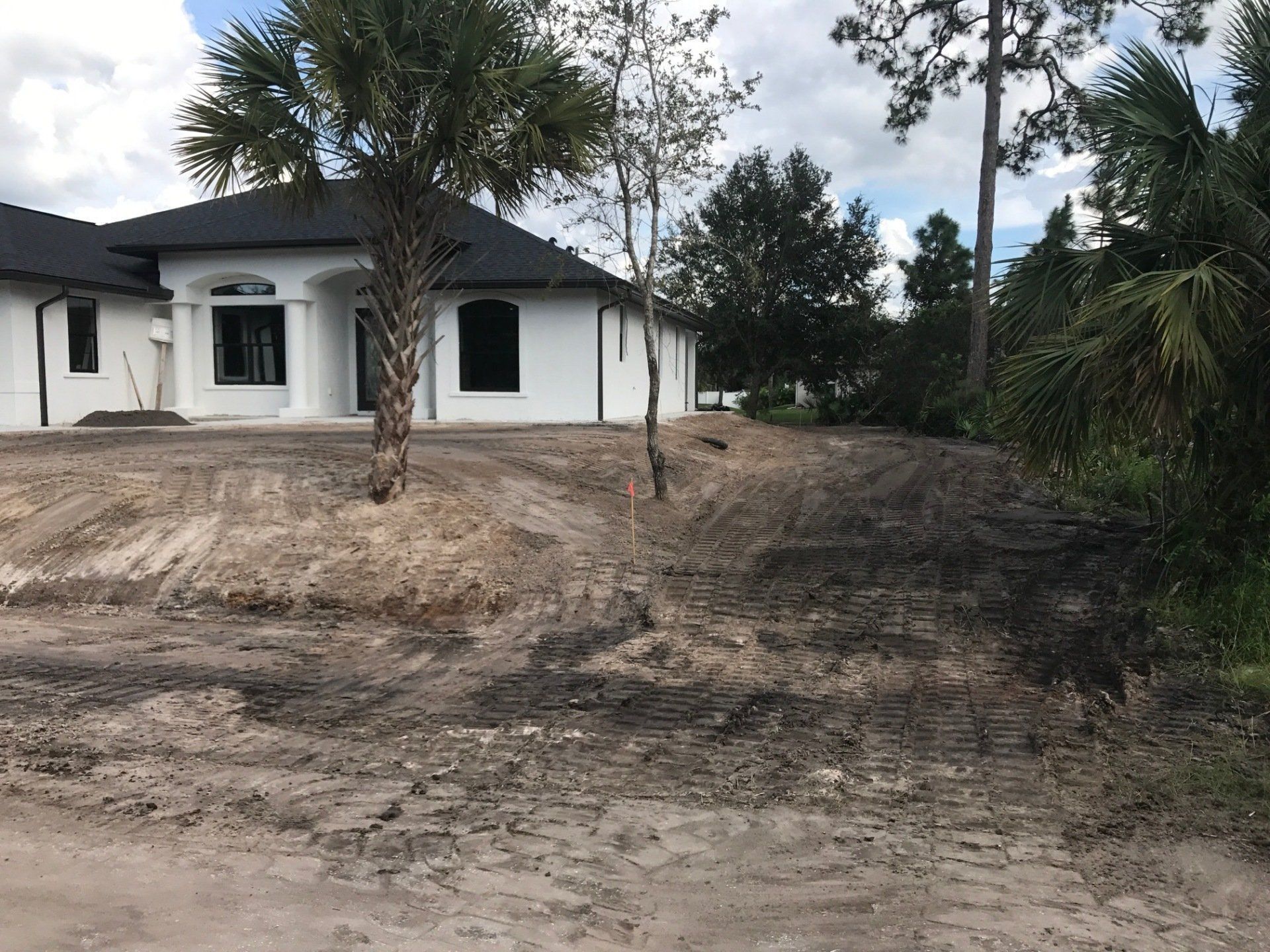 White house with black roof, set on a graded dirt lot, with a few trees and a cloudy sky.