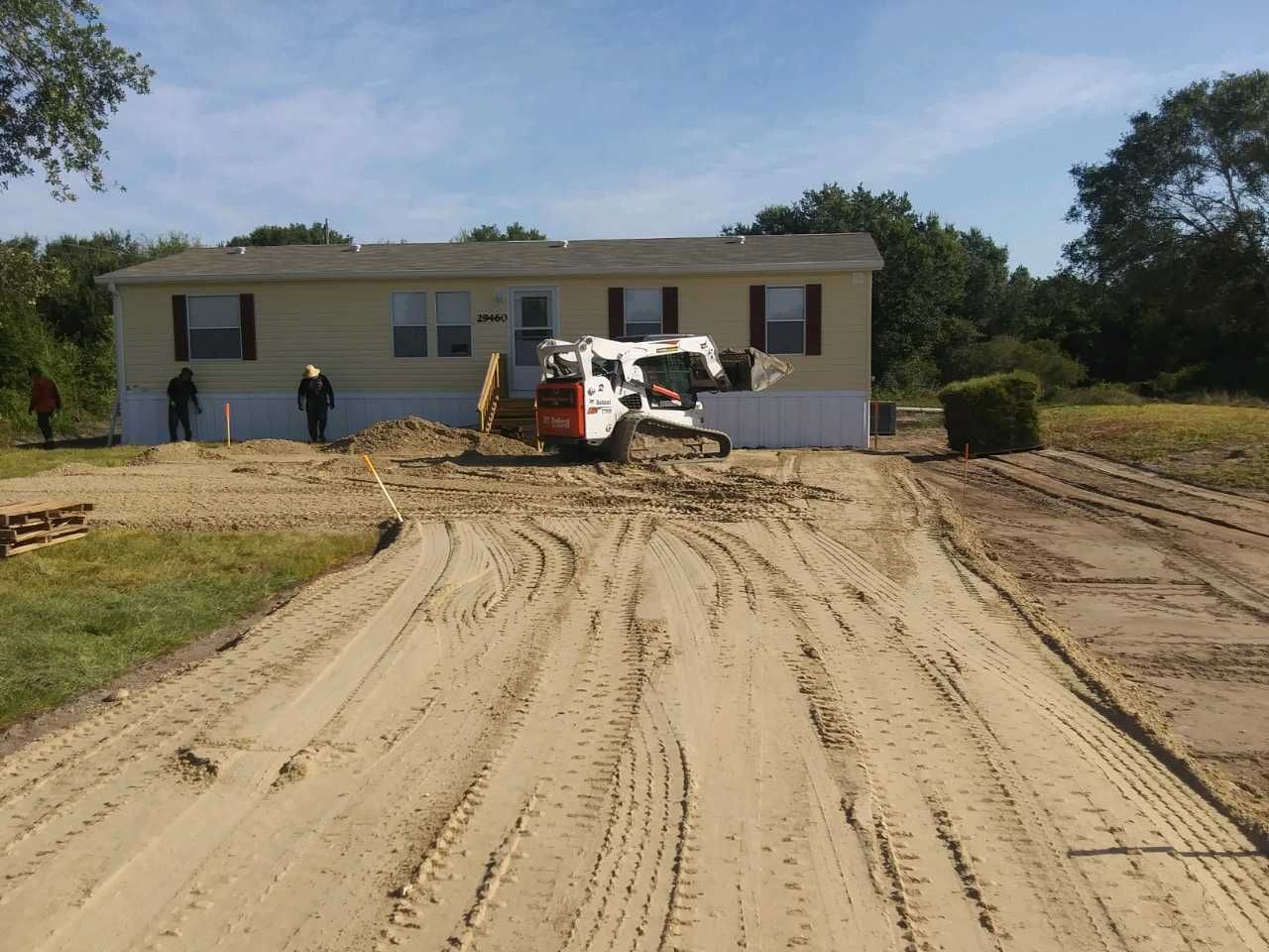 Bobcat and workers grading the sandy driveway of a single-story beige house under a blue sky.