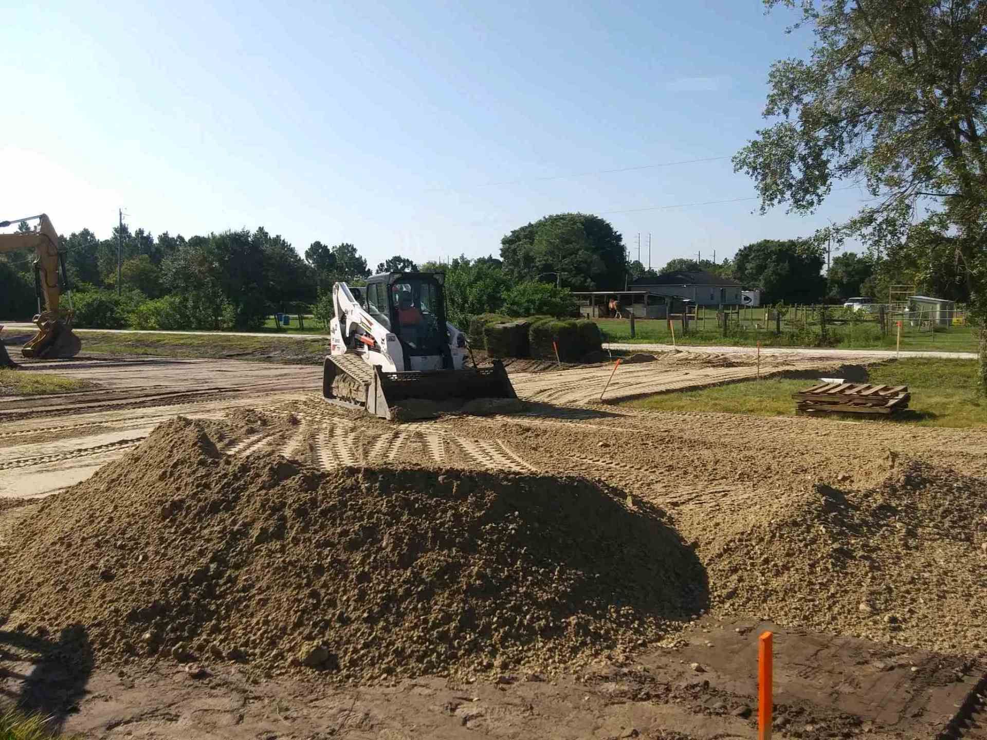 A small construction vehicle on a dirt lot, moving piles of dirt, with trees and a blue sky in the background.