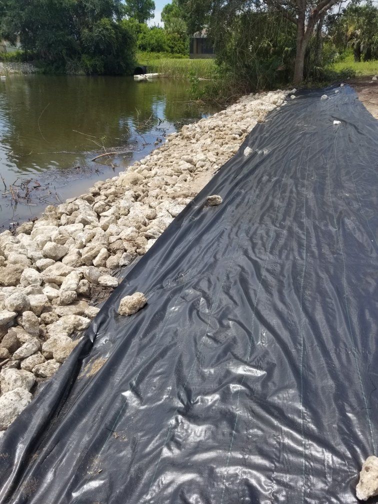 Riprap and black plastic on a shoreline to prevent erosion; water and trees in the background.