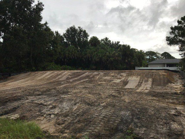 Dirt-covered land cleared for construction; trees and a house in the background under a cloudy sky.
