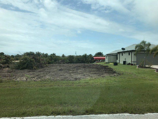Cleared lot with dark soil in front of a light-colored house under a partly cloudy sky.