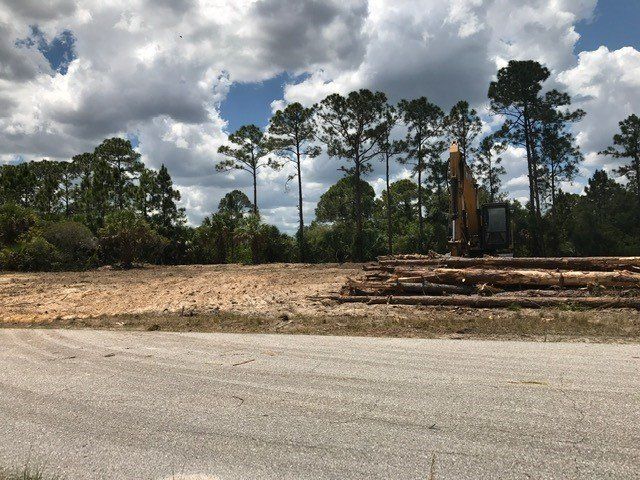 Clearing land with an excavator, logs piled up, trees in background, cloudy sky.