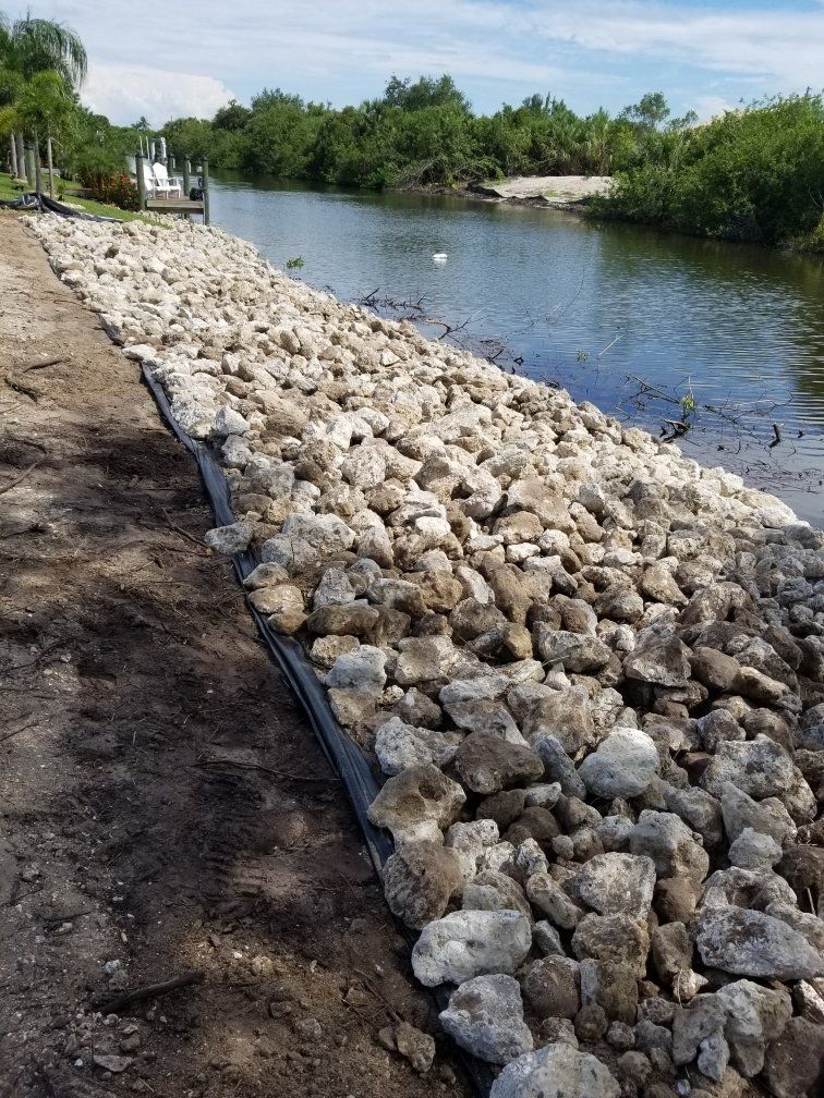 Rocks lining a canal bank. Light-colored rocks, dark mesh, brown soil, and water with trees in the background.