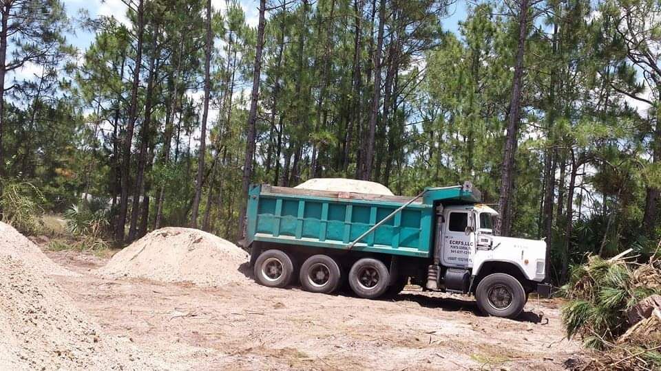 Teal dump truck unloading sand on a dirt lot, surrounded by trees.