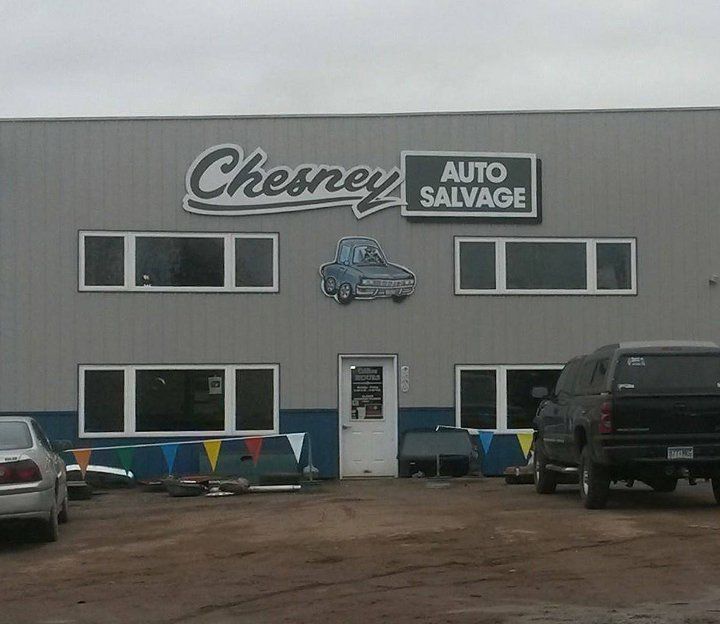 Chesney Auto Salvage building with sign, car graphic, and parked vehicles. Grey exterior, cloudy sky.