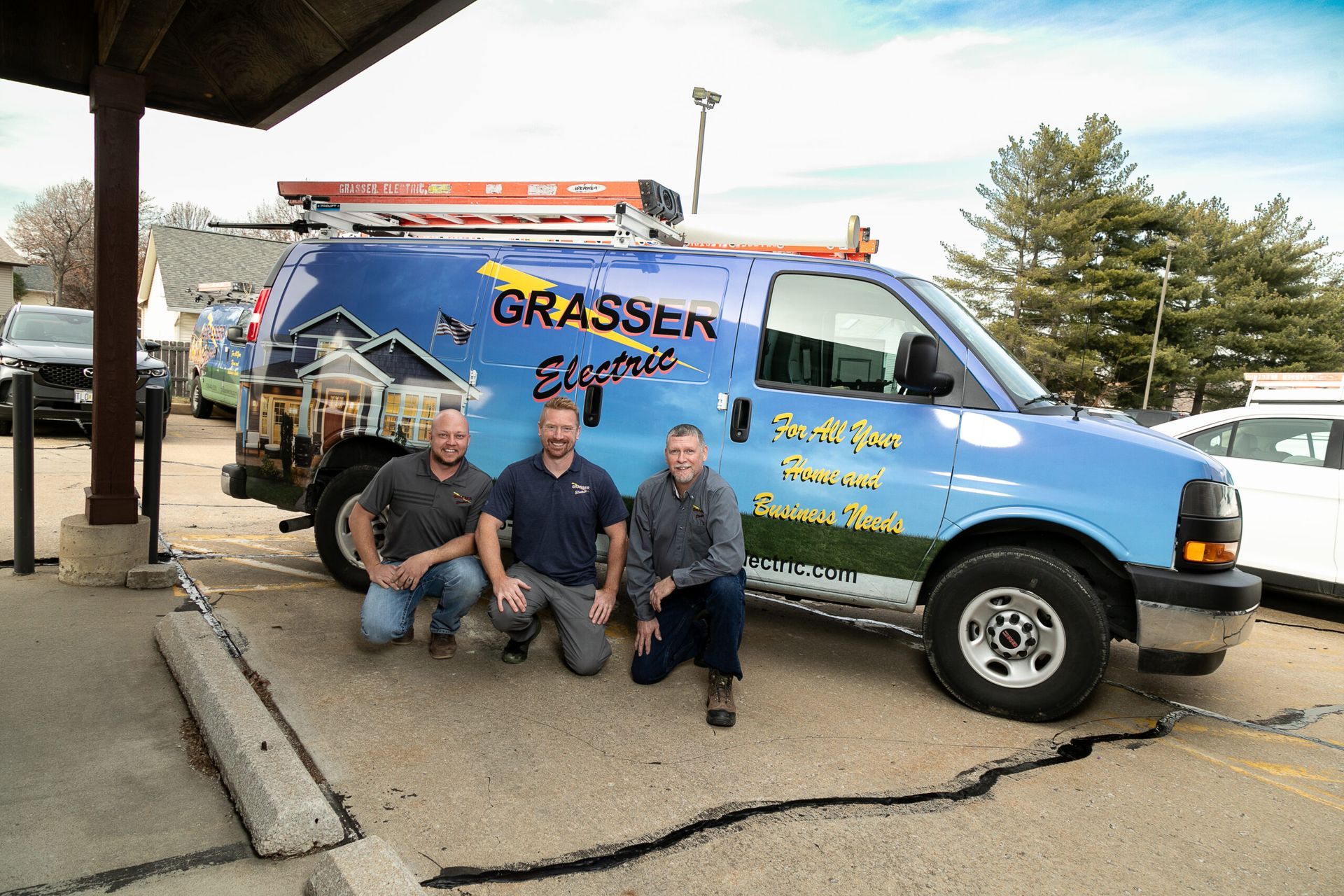 Three men kneel beside a blue Grasshopper service van under a covered driveway.
