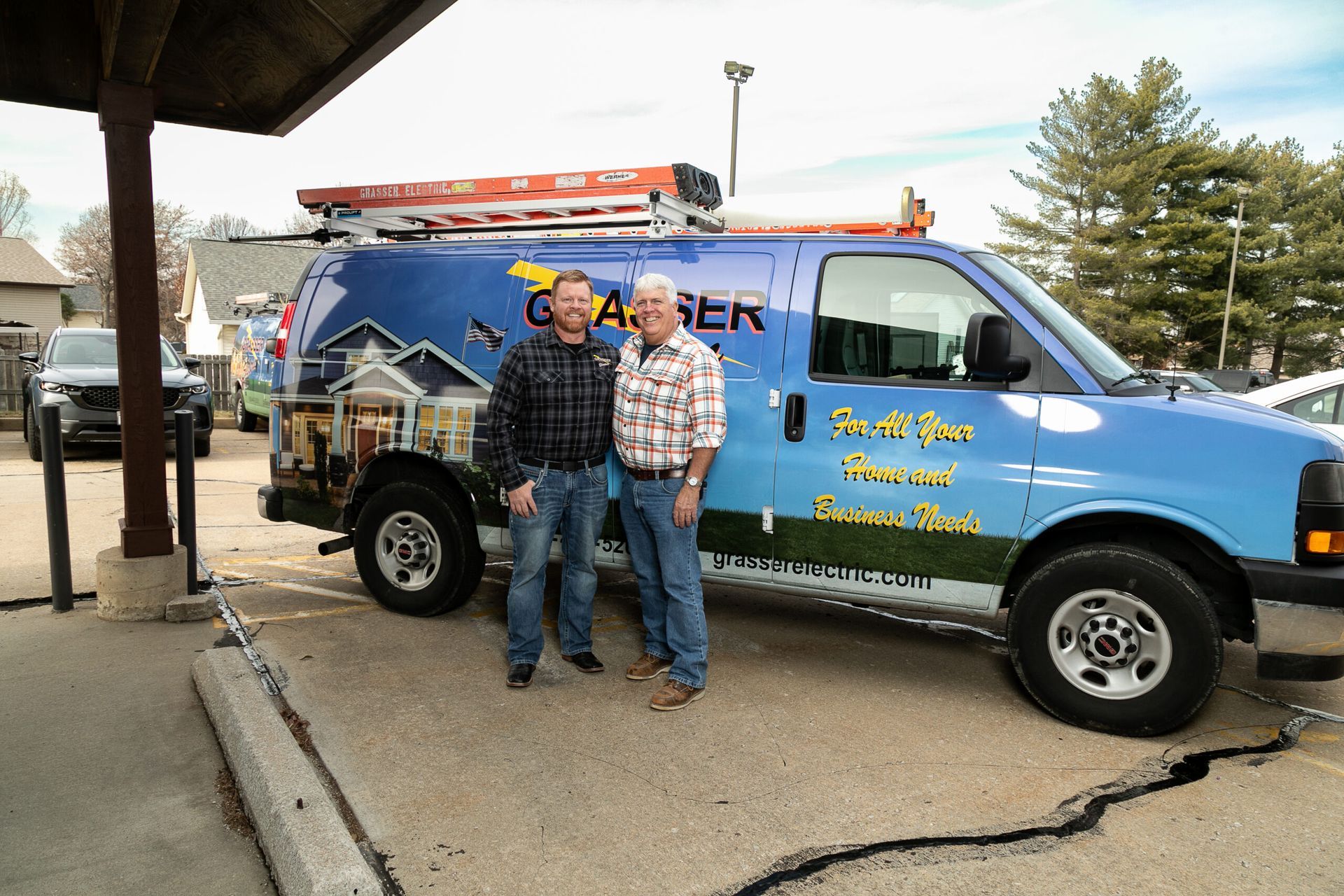Two men posing beside a blue service van with a ladder rack outdoors.