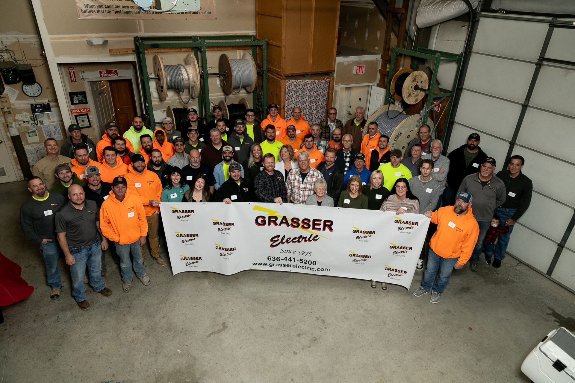 Group of workers posing in a warehouse, holding a “Grasser” banner and wearing safety vests and orange shirts