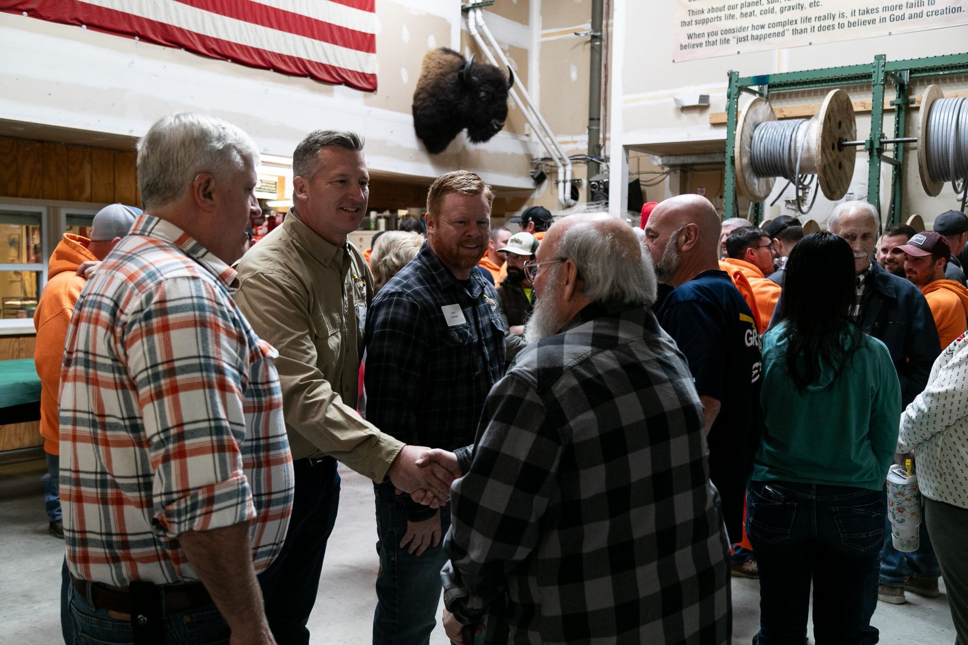 People shaking hands in a workshop, with machinery and an American flag in the background