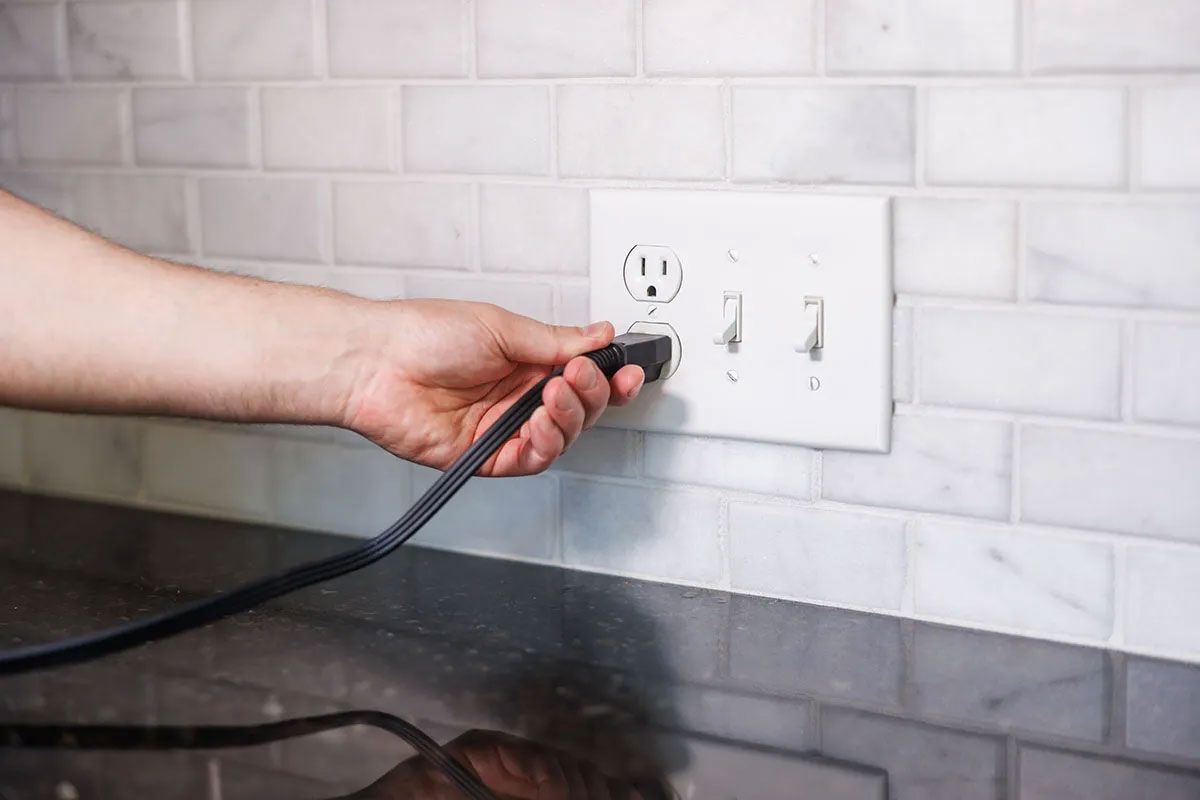 Hand plugging a black cord into a white wall outlet on a tiled kitchen backsplash