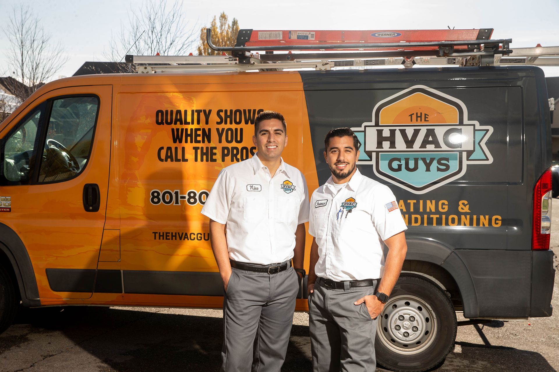Two HVAC technicians stand in front of their van. The orange and gray van has 
