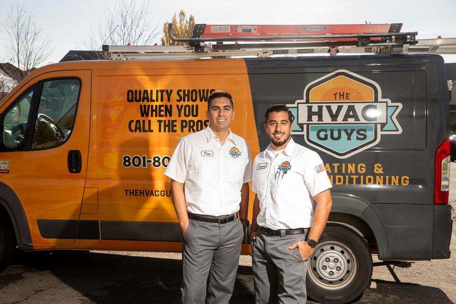 Two HVAC technicians stand in front of their van. The orange and gray van has 