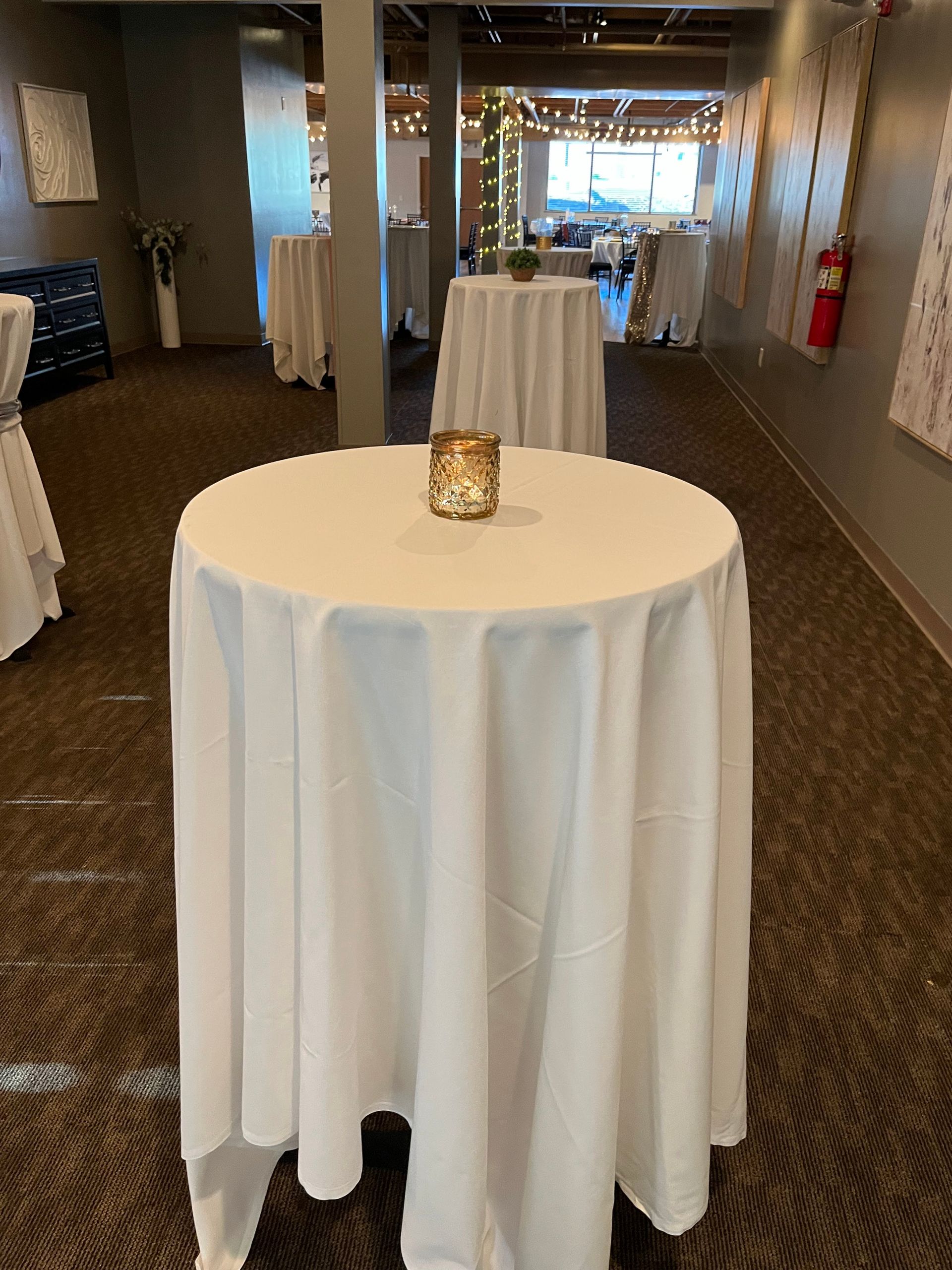 Cocktail tables with white linens in a hallway hallway leading to a bright event space