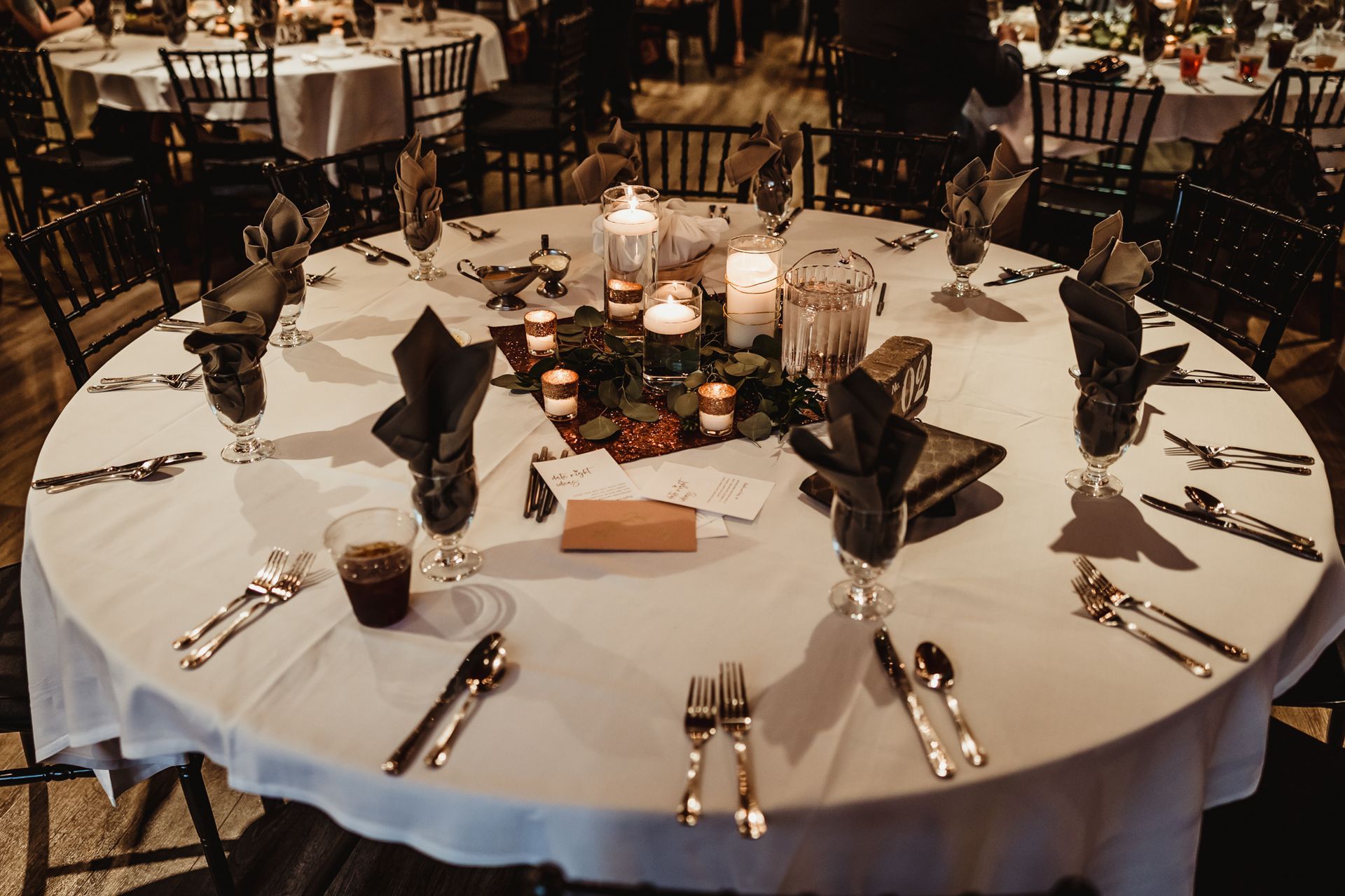 Elegant round banquet table set with white tablecloth, candles, folded black napkins, and silverware in a dimly lit hall