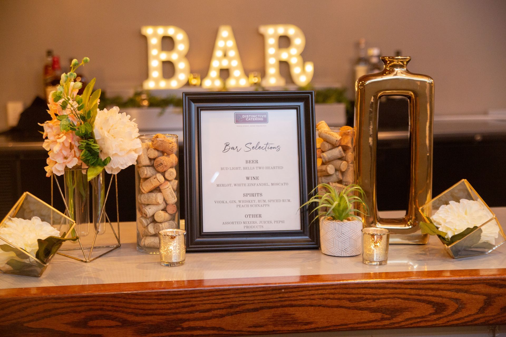 Bar display with framed sign, flowers, candles, jars, and lit marquee letters on a wooden table.