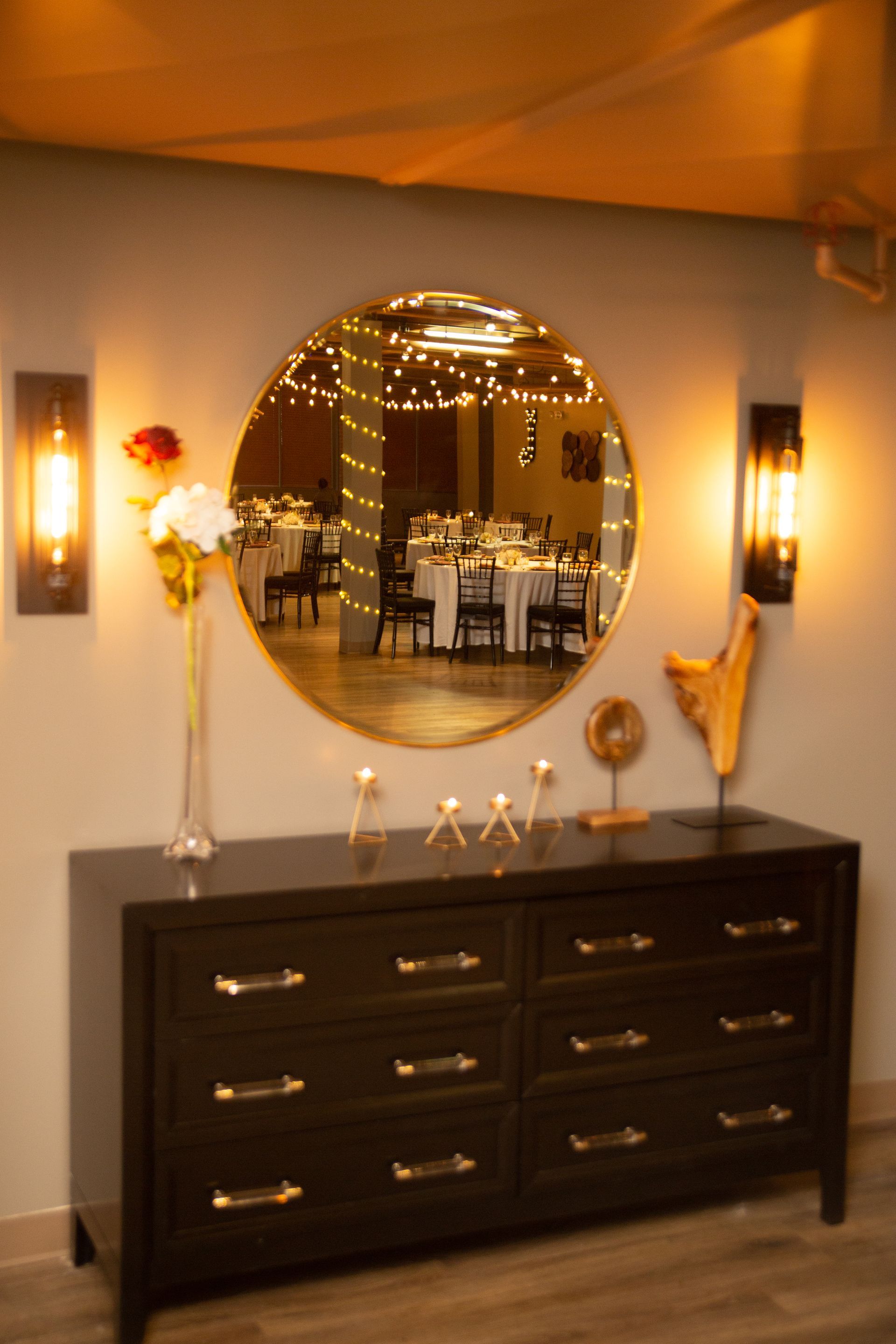 Dark wood dresser with round mirror, wall sconces, and decorative vases in a warmly lit room