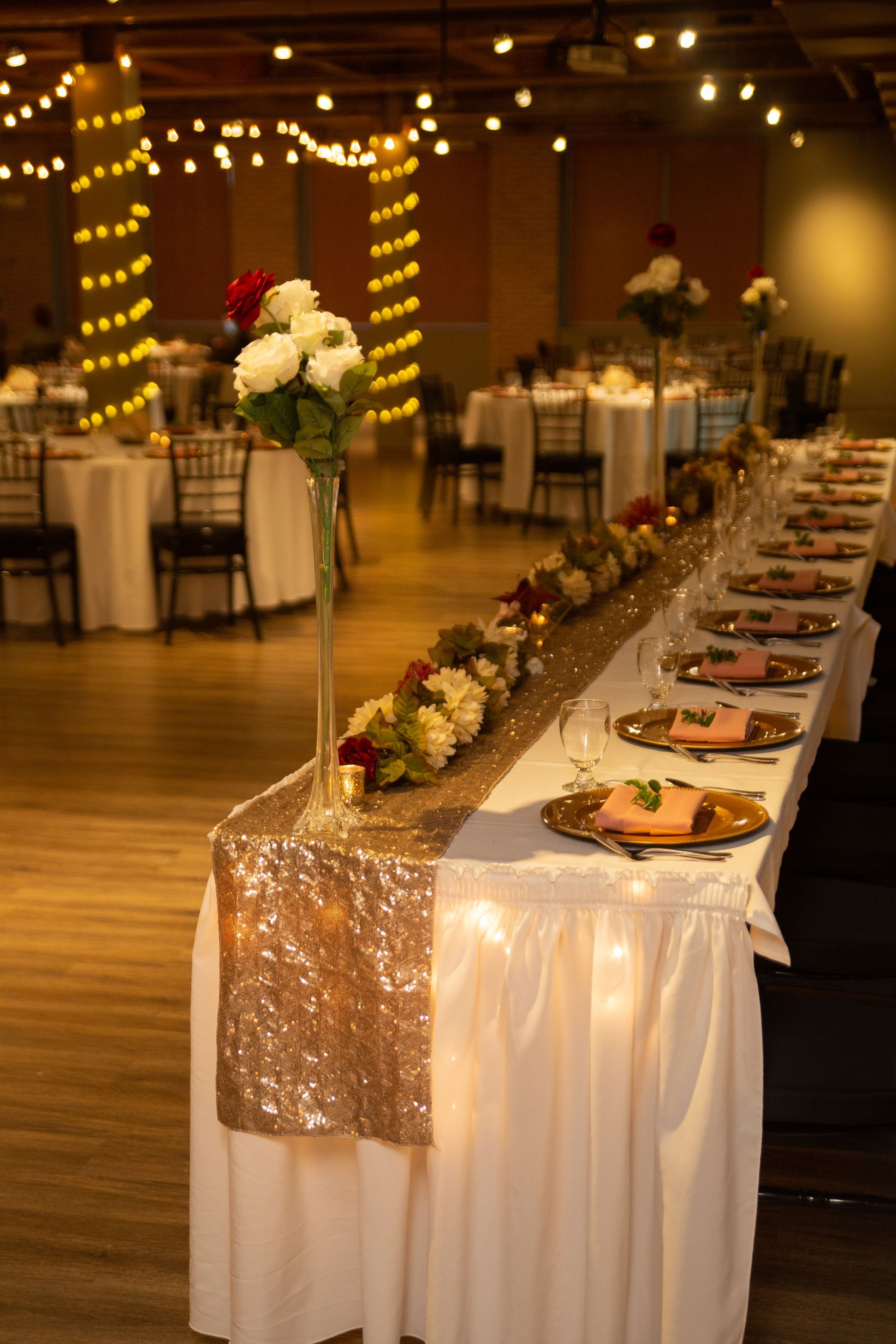 Long banquet table with white linens and floral centerpieces in a warmly lit event hall