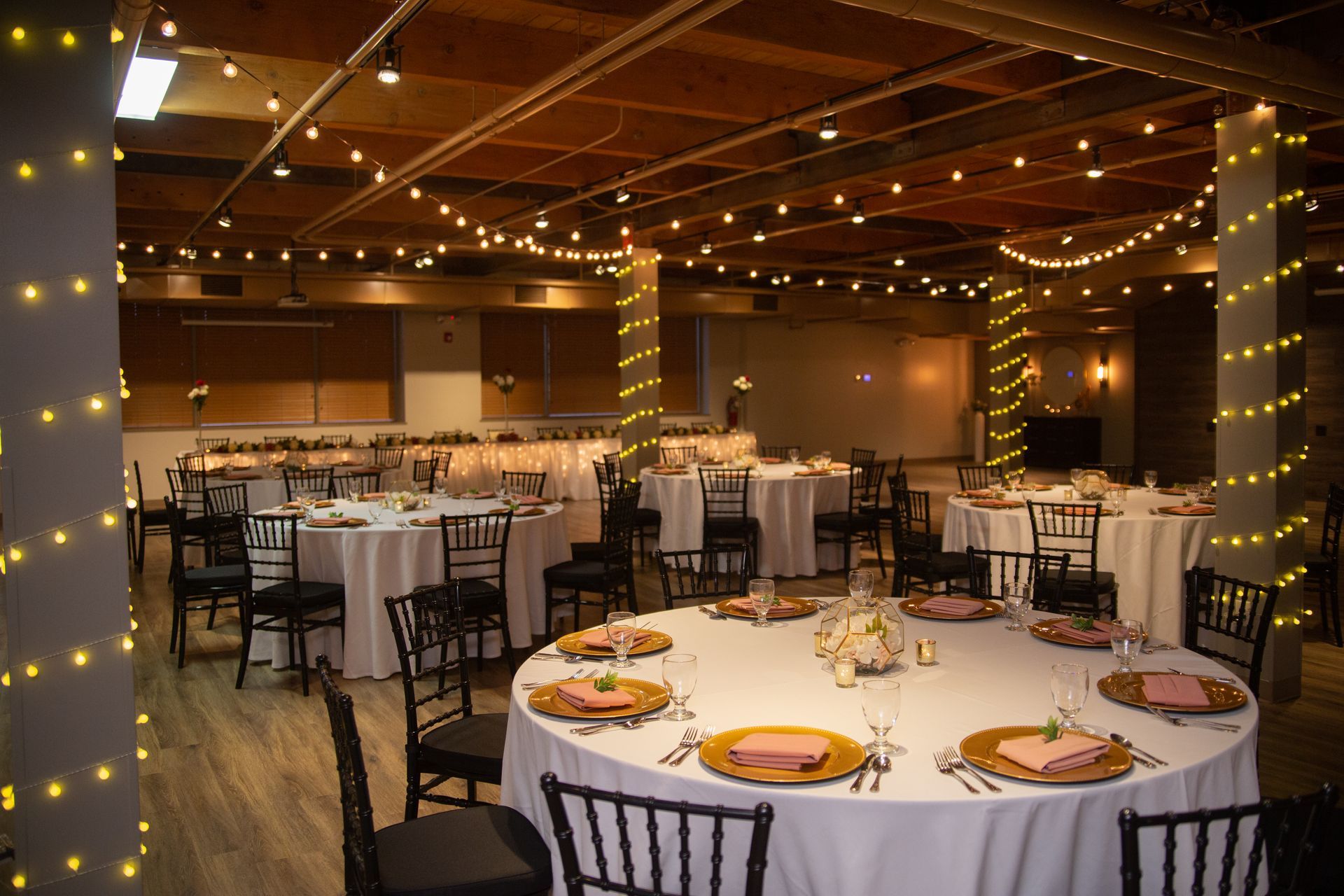Elegant banquet hall with round white-tablecloth tables, black chairs, and warm string lights overhead