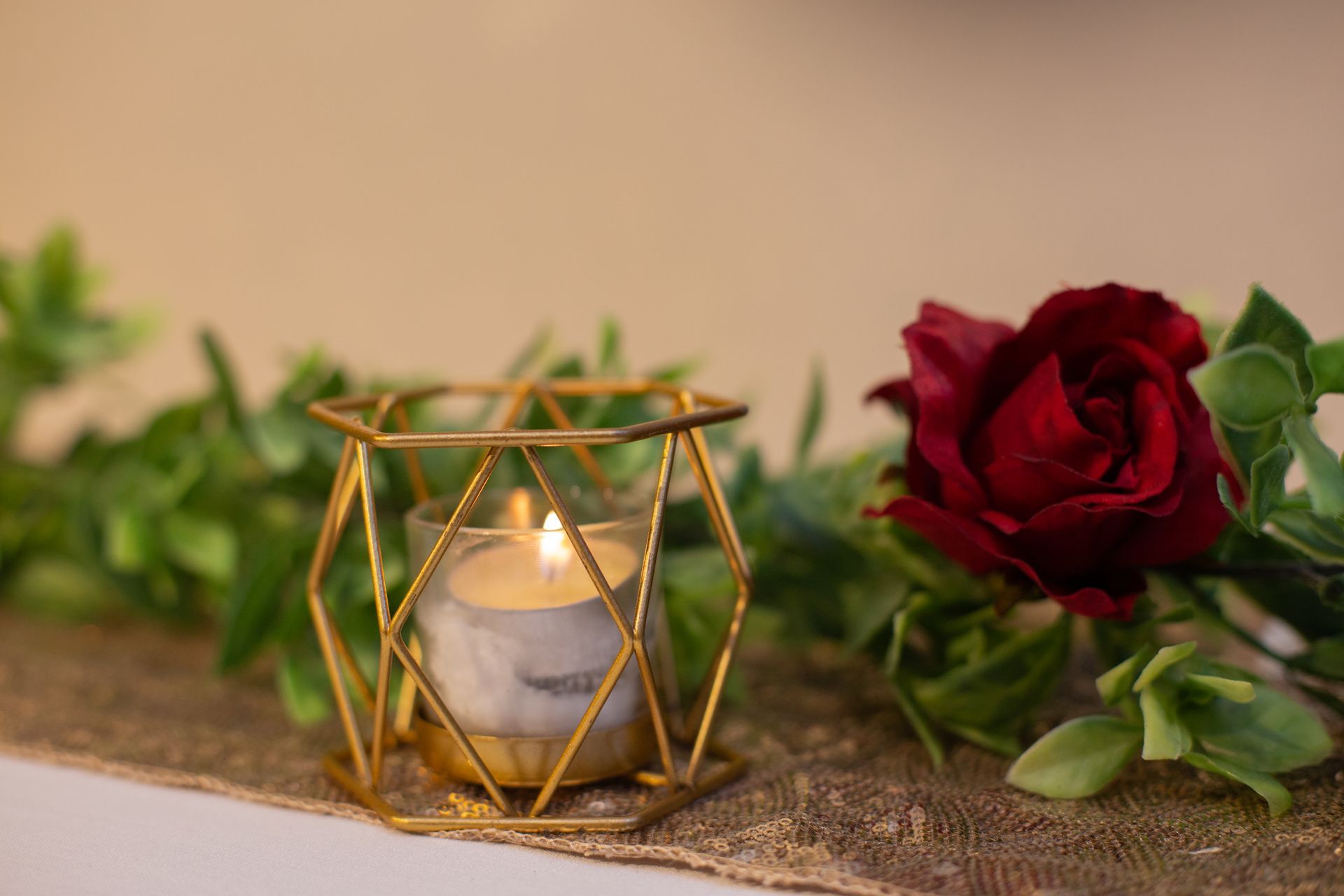 Lit tealight in gold geometric holder beside red rose on burlap table runner