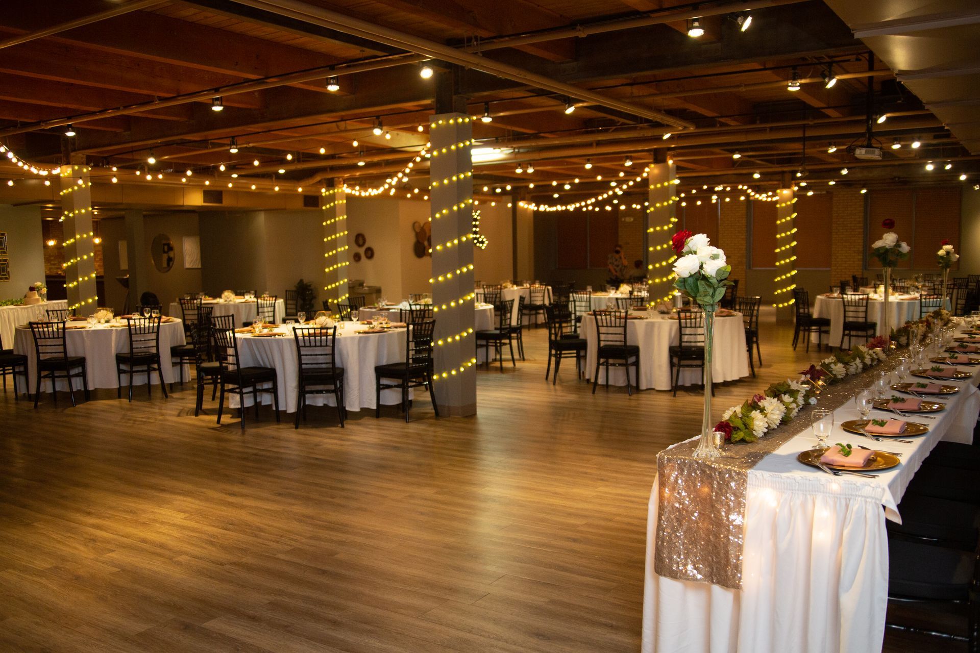 Decorated banquet hall with string lights, round tables, and a buffet setup on polished wood flooring