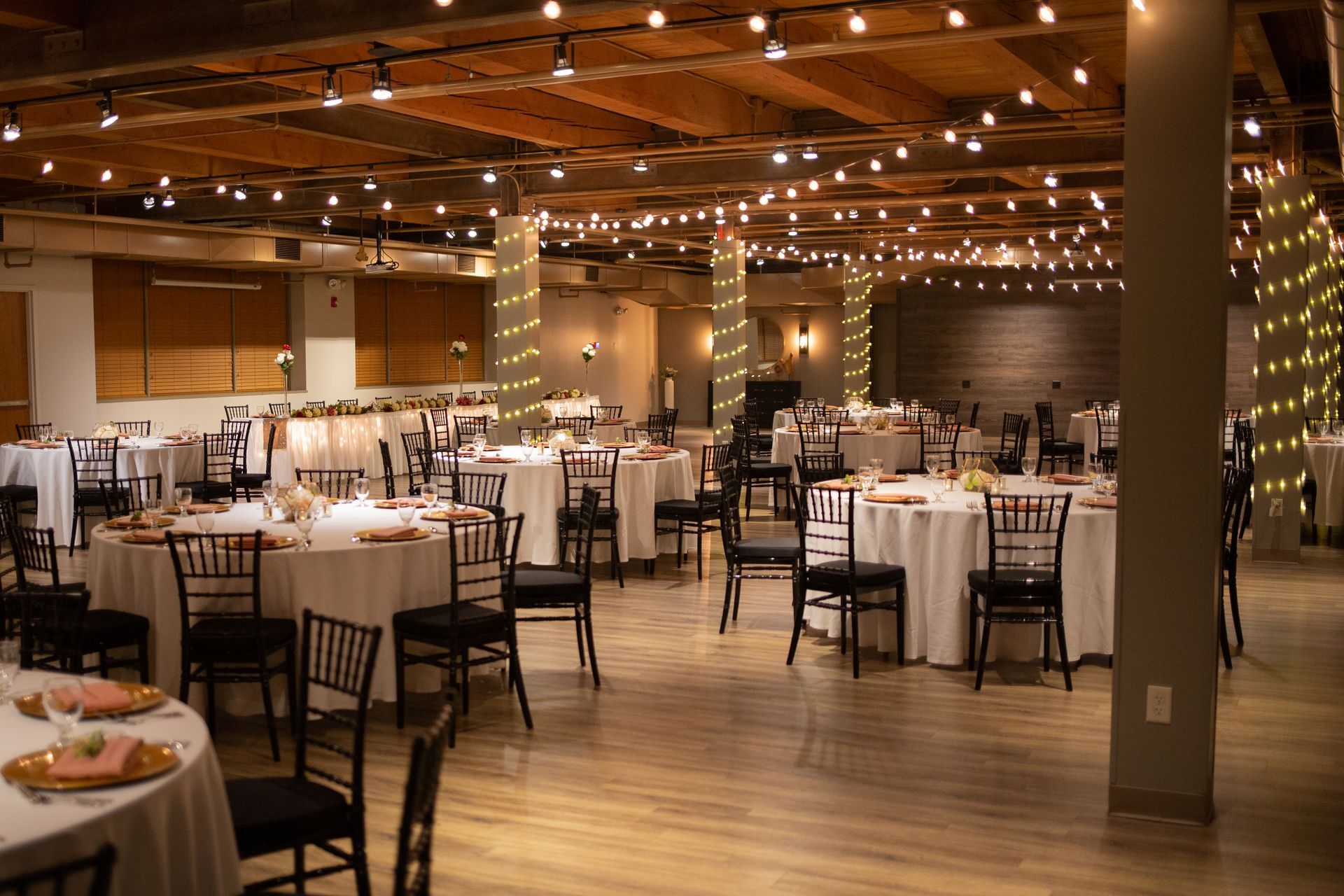 Empty banquet hall with round tables, black chairs, and warm string lights under a wooden ceiling